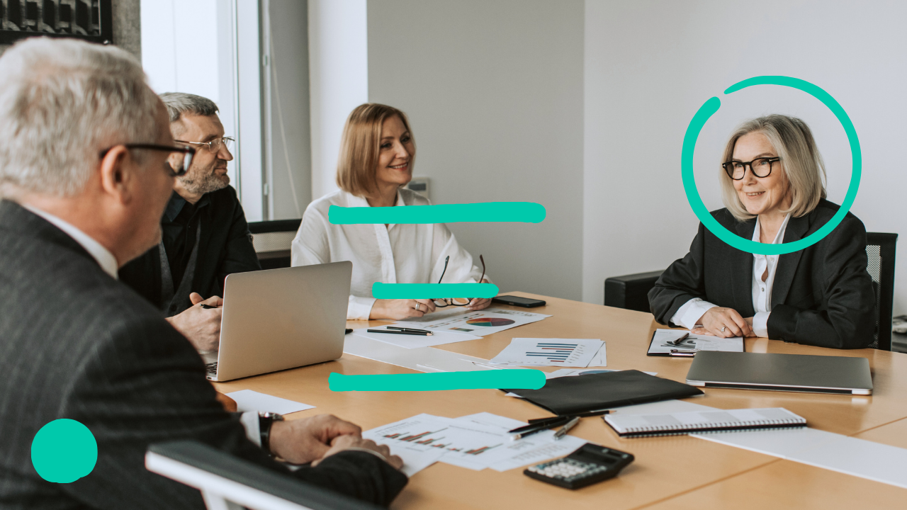 Four people in business attire are seated around a conference table with papers and a laptop scattered across it. They appear to be in a meeting.
