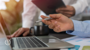 A close-up of a person's hand holding a pen over a laptop, with another person holding a tablet in the background.