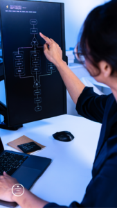 A person points at a flowchart on a computer monitor while working on a laptop with code displayed on the screen.
