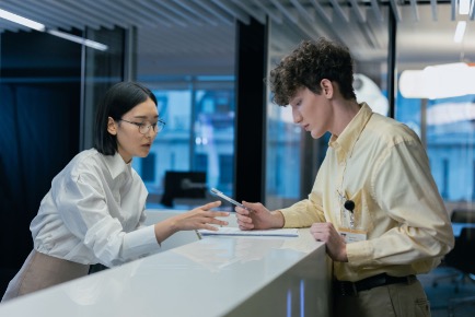 Photograph shows two coworkers discussing a document beside a modern office reception desk. Glass walls, blue ambient lighting, white counter, and visible identification badges suggest a professional workplace consultation.